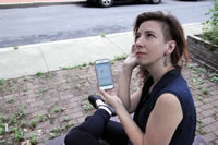 Stephanie Loveless sitting on a bench in a downtown area. She is holding a smart-phone running the Listening Here: Albany app. The instruction is "Listen for a sound you've never heard before." and it is accompanied by a picture of an ear with a soundwave going into it.